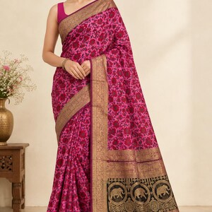 Puede incluir: Un sari de color magenta vibrante con un patrón floral oscuro que se repite por toda la tela. La prenda incluye un borde dorado ancho y ornamentado con detalles intrincados y un panel negro prominente en el extremo con motivos de elefantes dorados.