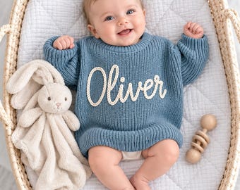 Op de afbeelding: Een blauwe, geribbelde gebreide trui met de naam Oliver in witte schrijfletters op de borst. De trui wordt gedragen door een baby die in een gevlochten mand ligt naast een beige pluche konijn en een houten rammelaar.