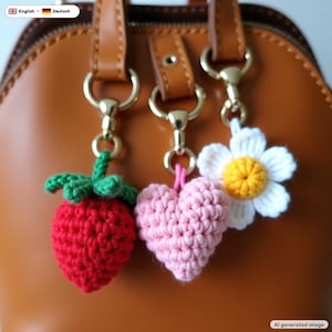 May include: Three crochet charms hanging from gold-toned metal clasps attached to a brown leather bag. The charms include a red strawberry with green leaves, a light pink heart, and a white flower with a yellow center.