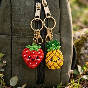 Può includere: Due portachiavi di perline attaccati ad anelli e chiusure in metallo dorato. Uno è una fragola rossa con foglie verdi e un viso sorridente. L'altro è un ananas giallo con foglie verdi e un viso sorridente, entrambi appesi a una borsa verde scuro.