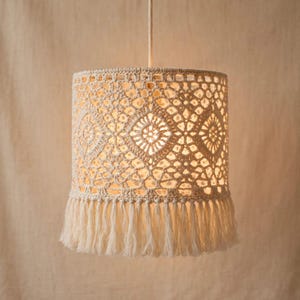 May include: A cylindrical pendant lamp shade crafted from off-white macrame cord featuring an intricate geometric diamond pattern. The shade is suspended by a single cord and finished with a long, straight fringe along the bottom edge.