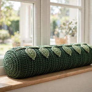 May include: A cylindrical bolster cushion crocheted in a deep forest green yarn. The cushion features a decorative vine motif running along the top, consisting of several light sage green crocheted leaves attached to a central stem.