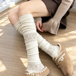 Puede incluir: Calentadores de piernas acanalados de color crema con un sutil patrón de corazones marrones en los puños y la parte central, combinados con zapatos sin cordones de ante beige con ribete blanco decorativo y hebilla metálica dorada.