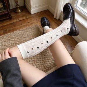 May include: Off-white knee-high socks featuring a delicate, repeating open-knit pattern and small, scattered black and brown heart-shaped embellishments, paired with dark brown leather loafers with a classic penny slot design.