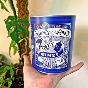 May include: A blue metal container featuring the words "Wibbly Wobbly Timey Wimey Stuff" in white, with clock and police box illustrations. The container is held in a hand, with a plant in the background.