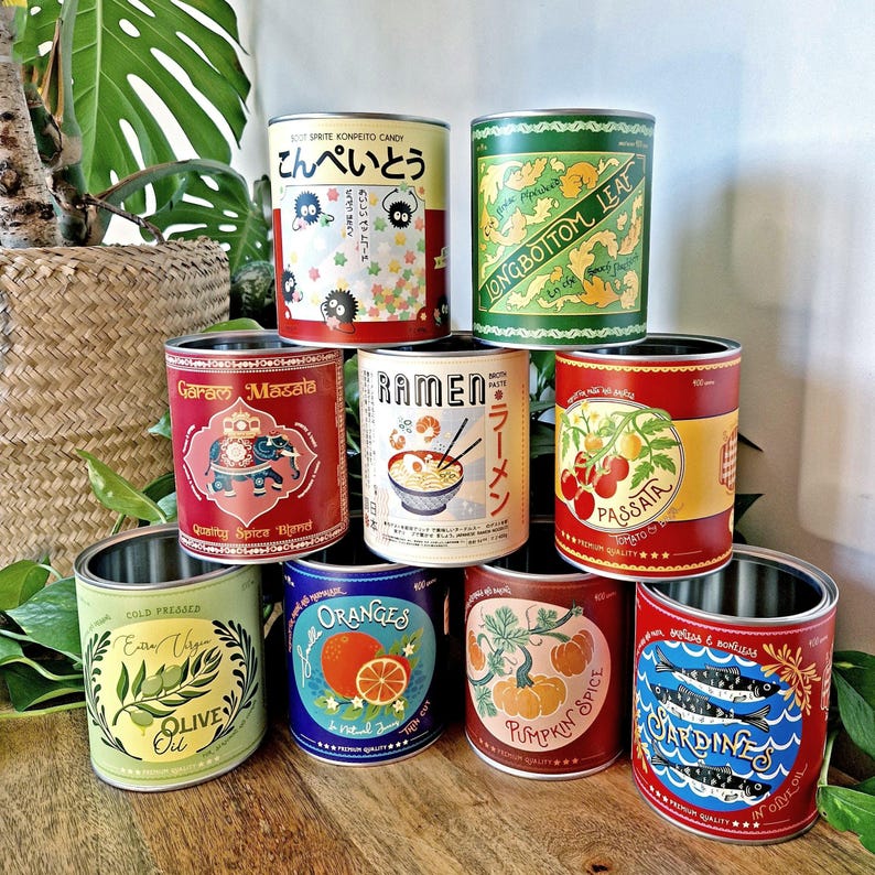 May include: A collection of decorative metal cans with various labels. The labels feature text and illustrations, including "Garam Masala," "Ramen," "Passata," "Olive Oil," "Oranges," "Pumpkin Spice," and "Sardines." The cans are stacked on a wooden surface.