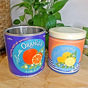 May include: Two cylindrical metal storage tins. The left tin is purple with a label reading Seville Oranges, featuring a whole orange and a slice, marked 400g. The right tin is orange with a cream lid, labelled Preserved Lemons with citrus graphics.