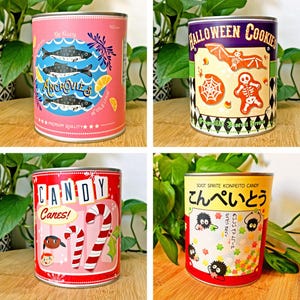 May include: Four decorative tin cans with colorful labels. One pink can features anchovies, another has Halloween cookies, a third displays candy canes, and the last one showcases Japanese candy with text in Japanese.