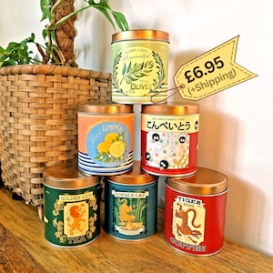 May include: Six cylindrical metal tins with copper-coloured lids, featuring various labels: Rosemary & Garlic Marinated Pitted Olives, Lemon, Soot Sprite Konpeito Candy, Golden Lion Tea, Jungle Cat Sugar, and Tiger Brand Coffee with colourful graphics.