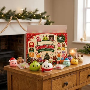 Op de afbeelding: Een rode, rechthoekige Dumpling-adventskalenderdoos met 24 verrassingen om te ontdekken, versierd met feestelijke illustraties. Verschillende kleine, kleurrijke, lachende knoedelfiguurtjes staan voor de doos op een houten oppervlak.