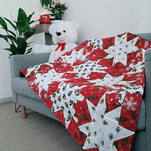Pode incluir: Uma manta acolchoada com um padrão repetitivo de estrelas em tecido branco sobre um fundo vermelho. O tecido é estampado com motivos festivos, incluindo árvores de Natal verdes, bagas de azevinho e flocos de neve brancos no design.