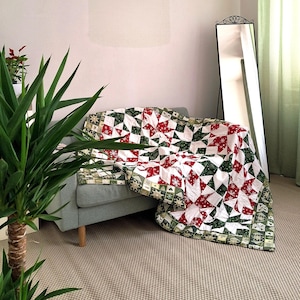 Peut inclure: Un quilt en patchwork présentant un motif d'étoiles répétitif en rouge, blanc et vert foncé. Le tissu affiche des motifs festifs incluant des flocons de neige, des sapins et des cœurs. Le quilt est drapé sur un canapé gris clair.