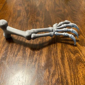 Może przedstawiać: Szary, wydrukowany w technologii 3D model szkieletu ręki i przedramienia. Obiekt posiada zaokrąglony staw w miejscu ramienia, strukturę przedramienia oraz szczegółowo odwzorowaną dłoń z pięcioma palcami zakończonymi ostrymi końcówkami.