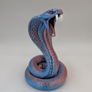 May include: A coiled cobra figurine featuring a metallic blue and purple gradient finish. The snake has an open mouth revealing a red interior, sharp white fangs, and piercing red eyes, with detailed scales covering its entire body.