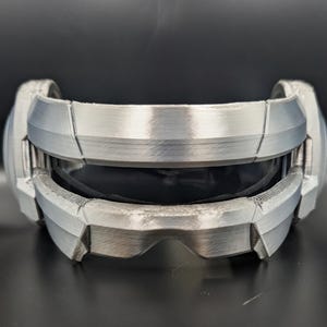 May include: A silver-colored visor with a metallic finish and a dark, tinted lens. The frame features a segmented, angular design with visible horizontal lines across the surface, creating a structured, futuristic appearance for the eyewear.