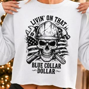 Puede incluir: Una sudadera blanca con un gráfico negro de una calavera con casco de obra frente a una bandera estadounidense. Herramientas cruzadas, una llave inglesa y un martillo, están detrás de la calavera. El texto dice LIVIN' ON THAT BLUE COLLAR DOLLAR.