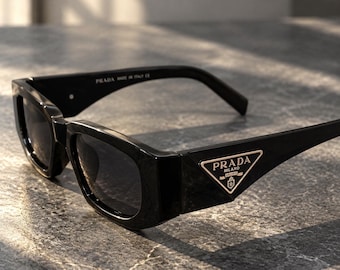 May include: Black rectangular sunglasses with dark lenses and thick frames. A gold-tone triangular Prada Milano logo plaque is featured on the temple. The inner arm displays the text Prada Made in Italy CE in white lettering.
