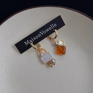 Puede incluir: Un par de pendientes de aro dorados sujetos a una tarjeta negra con el texto MaisonVowelle. Un pendiente tiene una cuenta rectangular blanca y una perla pequeña, el otro una cuenta color ámbar y una perla pequeña.