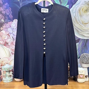 May include: A dark navy blue long-sleeve jacket featuring a round neckline and a vertical row of ten silver-toned buttons down the center front. The jacket has a straight hemline with a split opening at the bottom and a label reading DANI MAY.