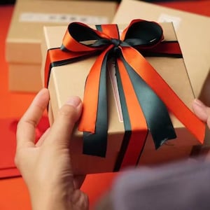 Op de afbeelding: Een vierkante kartonnen geschenkdoos vastgebonden met een decoratief lint met afwisselende oranje en zwarte satijnen strepen. De doos is lichtbruin en heeft een strik in het midden, met andere soortgelijke dozen op de achtergrond.