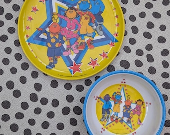 Puede incluir: Un plato amarillo y un cuenco blanco con borde azul, ambos con una ilustración gráfica de cuatro personajes de dibujos animados de pie dentro de una estrella azul rodeada de pequeñas estrellas rojas. Los personajes llevan ropa colorida.