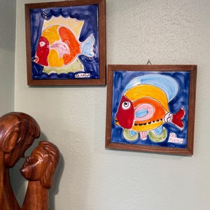 Könnte beinhalten: Zwei quadratische Keramikfliesen in Holzrahmen, die jeweils einen handbemalten Fisch in leuchtenden Rot-, Orange-, Gelb- und Blautönen vor einem tiefblauen Hintergrund zeigen. Jede Fliese ist in der Ecke mit "La Musa" signiert.