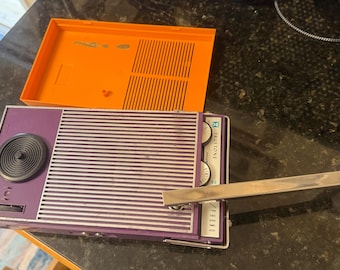 Può includere: Una radio portatile Truetone viola e argento con altoparlante, maniglia in metallo argentato e due manopole di controllo. Il dispositivo presenta un coperchio posteriore in plastica arancione brillante staccato con una griglia.