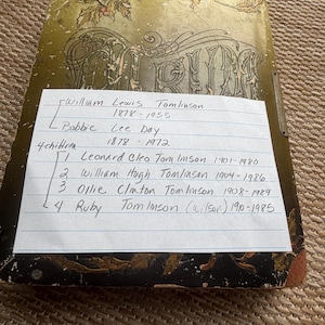 Könnte beinhalten: Ein antikes Fotoalbum mit einem abgenutzten gold-schwarzen Einband, der florale Illustrationen und das Wort Album zeigt. Auf dem Album liegt ein liniertes Blatt Papier mit handschriftlichen genealogischen Notizen zu William Lewis Tomlinson, Bobbie Lee Day und deren vier Kindern mit Geburts- und Sterbejahr.