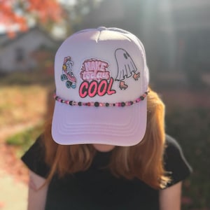 May include: A white trucker hat featuring a beaded trim along the brim and three patches on the front: a pink roller skate, the text "MAKE today COOL" in pink and black, and a white ghost wearing orange roller skates.