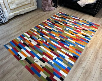 May include: A rectangular area rug constructed from individual rectangular patches of cowhide leather. The rug features a vibrant, multi-coloured pattern of red, orange, blue, green, black, and white segments, including some patches with black and white cowhide spots.