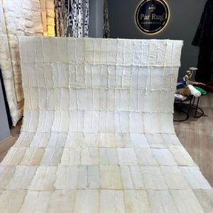 Op de afbeelding: Een groot, rechthoekig vloerkleed opgebouwd uit talloze gebroken witte en crèmekleurige rechthoekige leren stukken die in een rasterpatroon aan elkaar zijn genaaid. Het oppervlak heeft een harige textuur met subtiele kleurnuances.