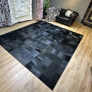 Op de afbeelding: Een groot, rechthoekig vloerkleed bestaande uit talloze vierkante en rechthoekige stukken zwart rundleer. Het kleed heeft een patchworkontwerp met variërende texturen en tinten zwart, wat zorgt voor een uniform en genuanceerd oppervlak.