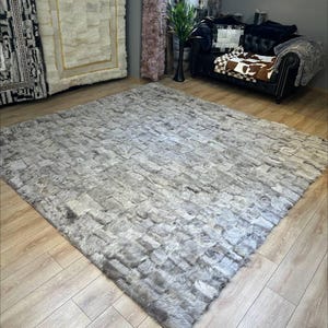 Könnte beinhalten: Ein großer, rechteckiger Teppich mit einem strukturierten Patchwork-Design in verschiedenen hellgrauen Farbtönen. Der Teppich besteht aus zahlreichen kleinen, rechteckigen Stücken aus fellartigem Material, die in einem Raster angeordnet sind.