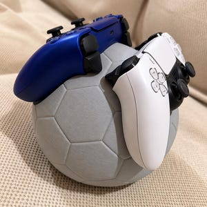 Op de afbeelding: Een grijze, bolvormige controllerstandaard in de vorm van een voetbal, met een getextureerd oppervlak. Een metallic blauwe draadloze gamecontroller rust aan de linkerkant, terwijl een witte draadloze gamecontroller aan de rechterkant staat.