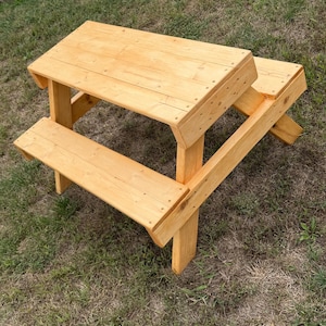 May include: A light-colored wooden picnic table featuring a rectangular tabletop and two attached benches on opposite sides. The structure is built with smooth, natural-toned wood planks joined together with visible screws, forming a sturdy frame.