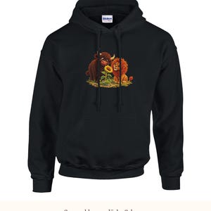K&ouml;nnte beinhalten: Ein schwarzer Kapuzenpullover mit einem Grafikdruck eines braunen Hochlandrindes und eines goldenen L&ouml;wen, die auf beiden Seiten einer gelben Sonnenblume sitzen. Der Hoodie verf&uuml;gt &uuml;ber eine K&auml;ngurutasche, lange &Auml;rmel und eine Kapuze.