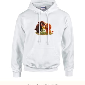 K&ouml;nnte beinhalten: Ein wei&szlig;er Kapuzenpullover mit einem Grafikdruck, der ein braunes Hochlandrind und einen L&ouml;wen zeigt, die zusammen neben einer gelben Sonnenblume sitzen. Der Hoodie hat eine K&auml;ngurutasche, lange &Auml;rmel und wei&szlig;e Kordelz&uuml;ge.