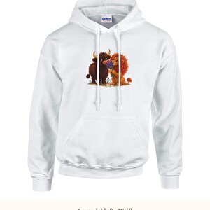 K&ouml;nnte beinhalten: Ein wei&szlig;er Kapuzenpullover mit einem Grafikdruck eines braunen Hochlandrindes und eines L&ouml;wen, der einen Strau&szlig; violetter Lavendelbl&uuml;ten h&auml;lt. Der Hoodie verf&uuml;gt &uuml;ber lange &Auml;rmel, eine K&auml;ngurutasche und wei&szlig;e Kordelz&uuml;ge.