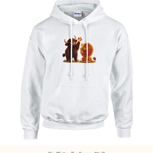 K&ouml;nnte beinhalten: Ein wei&szlig;er Kapuzenpullover mit einem Grafikdruck, der ein braunes Hochlandrind und einen goldenen L&ouml;wen zeigt, die zusammen auf gr&uuml;nem Gras sitzen, wobei das Rind nach einem fallenden orangefarbenen Herbstblatt greift. Mit langen &Auml;rmeln.