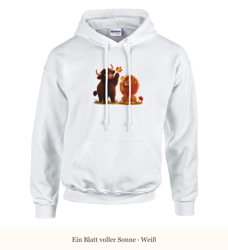 K&ouml;nnte beinhalten: Ein wei&szlig;er Kapuzenpullover mit einem Grafikdruck, der ein braunes Hochlandrind und einen goldenen L&ouml;wen zeigt, die zusammen auf gr&uuml;nem Gras sitzen, wobei das Rind nach einem fallenden orangefarbenen Herbstblatt greift. Mit langen &Auml;rmeln.