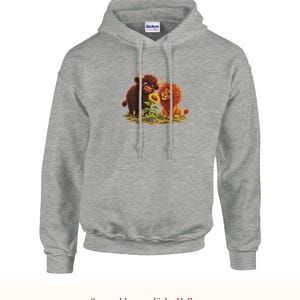 K&ouml;nnte beinhalten: Ein hellgrauer Kapuzenpullover mit einem Grafikdruck eines braunen Hochlandrindes und eines L&ouml;wenbabys, die zusammen neben einer gelben Sonnenblume sitzen. Das Kleidungsst&uuml;ck verf&uuml;gt &uuml;ber eine Kapuze mit Kordelzug und eine K&auml;ngurutasche.