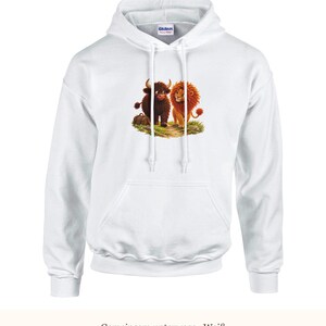 K&ouml;nnte beinhalten: Ein wei&szlig;er Kapuzenpullover mit einem Grafikdruck, der ein braunes Hochlandrind und einen goldenen L&ouml;wen zeigt, die zusammen auf einer gr&uuml;nen Grasfl&auml;che stehen. Der Hoodie hat eine K&auml;ngurutasche, lange &Auml;rmel und wei&szlig;e Kordelz&uuml;ge.