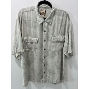May include: A light gray short-sleeve button-down shirt featuring a tonal leaf pattern. The shirt includes a pointed collar, two chest pockets with flaps, and a Tommy Bahama brand label on the inside neck. The fabric has a subtle texture.