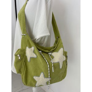 May include: A lime green corduroy hobo bag featuring three large, cream-colored fuzzy star appliqués. The bag includes a side pocket with a silver zipper, a wide shoulder strap, and a white drawstring cord with black speckles and plastic toggles.