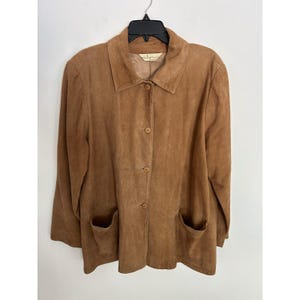 May include: A tan suede jacket featuring a classic collar, long sleeves, and a front button closure with four matching buttons. The garment includes two large patch pockets at the lower front and a visible interior label at the neckline.