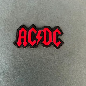 Könnte beinhalten: Ein Stoffaufnäher mit dem AC/DC-Logo in leuchtend roter Schrift und schwarzem Rand. Das Design enthält das ikonische Blitzsymbol zwischen den Buchstaben AC und DC, dargestellt in einer fetten Blockschrift auf einem schwarzen Hintergrund.