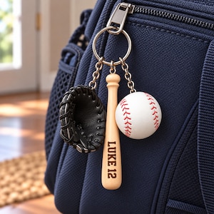 May include: A metal keychain featuring three charms: a black baseball glove, a light wood baseball bat engraved with the text LUKE 12, and a white baseball with red stitching, all attached to a silver ring clipped to a navy blue bag.