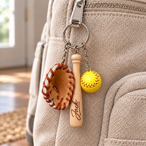May include: A metal keychain attached to a beige fabric bag, featuring three charms: a miniature brown leather baseball glove with white lacing, a light wood baseball bat engraved with the name Jack, and a small yellow softball with red stitching.