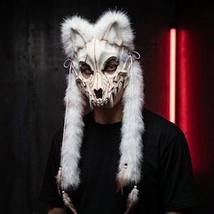May include: A white wolf skull mask featuring detailed bone-like textures with gold accents. The mask is adorned with long, fluffy white faux fur trim, white cord ties, and dangling brown feathers at the ends of the fur side panels.