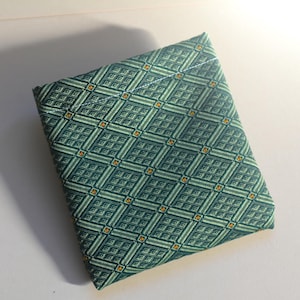 May include: A square fabric pouch featuring a teal and light green geometric diamond pattern with small orange accents. The textile has a structured appearance with a visible stitched seam along the top edge, creating a flat, envelope-style closure.